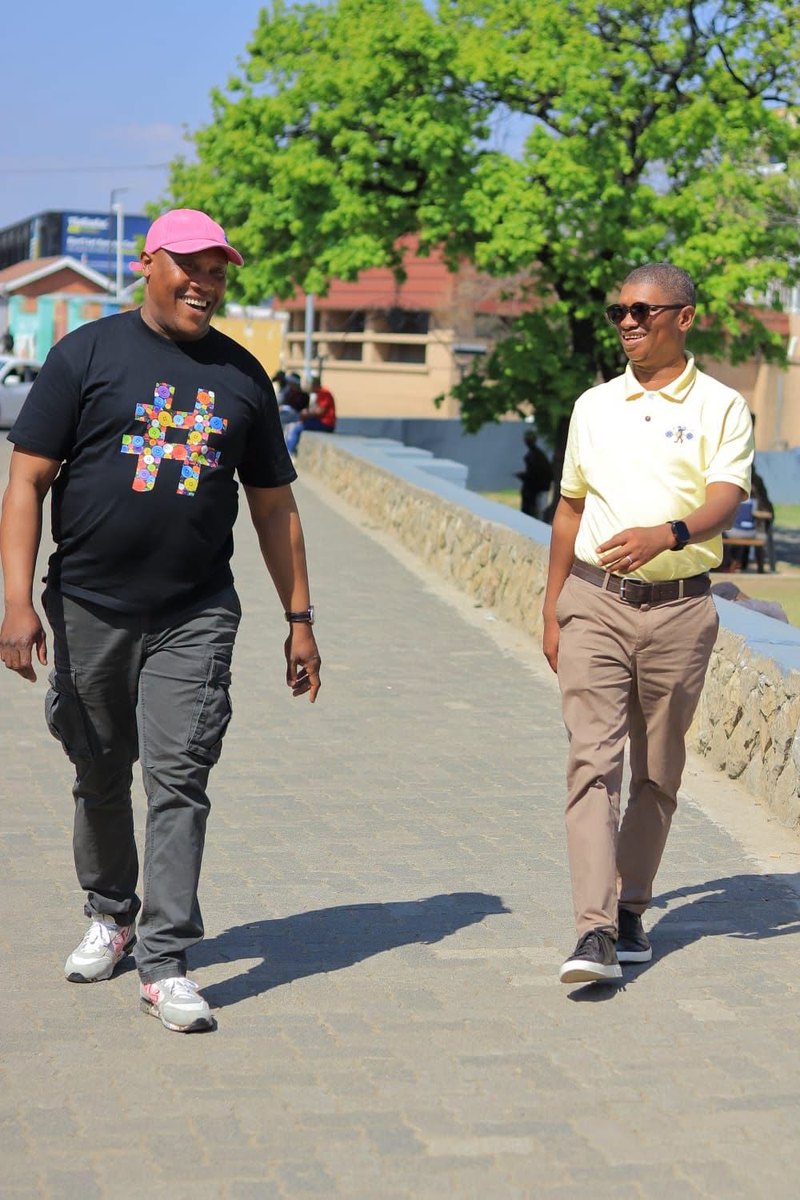 Two men walking, one in the black button hashtag tee and a pink bucket hat
